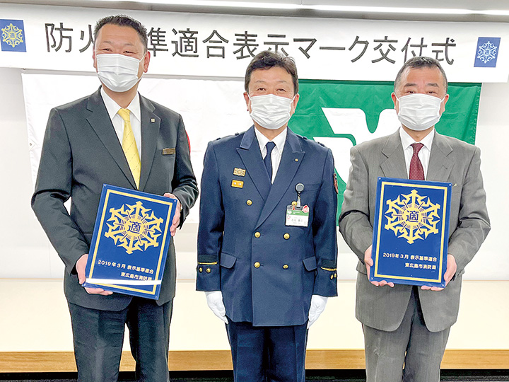 金の防火基準適合表示マークを手にする藤田副支配人（左）と坂田副支配人（右）。中央は石丸消防局長（撮影・林）