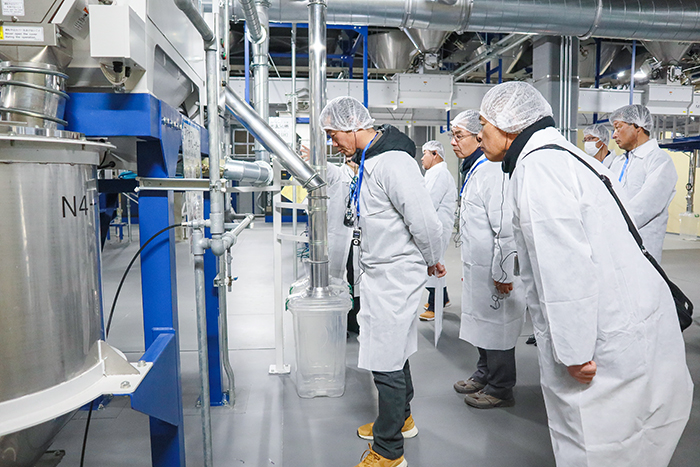 ネット帽と白衣を身に着けトランシーバーを装着して説明を聞きながら工場見学をする生産者ら(撮影・山北)