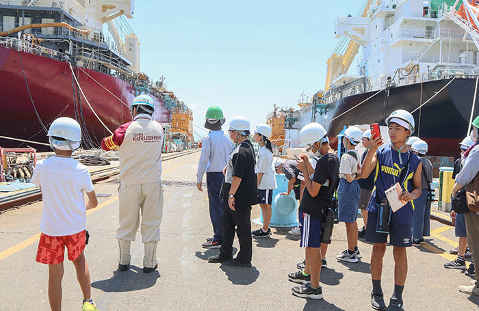 山野副工場長（左から２人目）から、船体の建造工程について説明を聞き、メモや撮影をしながら見学する子どもら